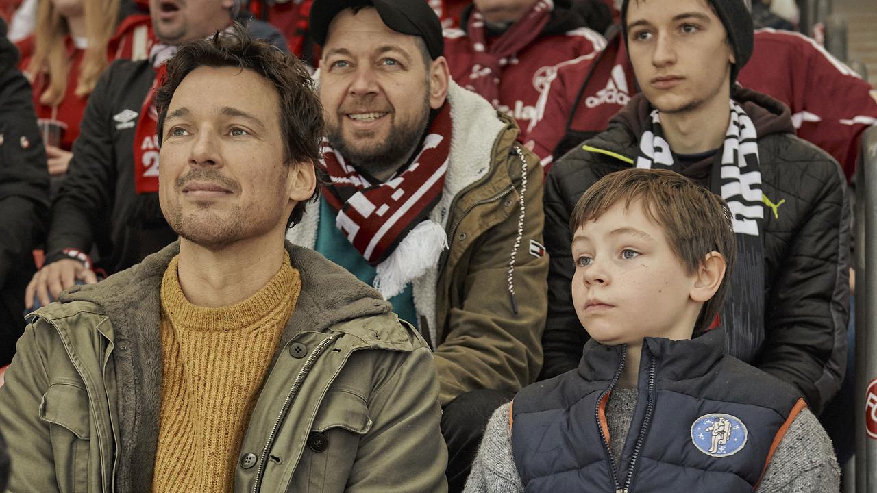 © ORF/Pro7/LEONINE Studios Mirco sitzt mit seinem Sohn Jason im Fußballstadion, hinter ihnen mehrere Personen in Fanbekleidung mit rot-weißen Schals