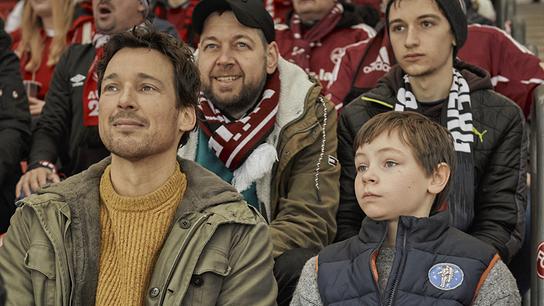 Mirco sitzt mit seinem Sohn Jason im Fußballstadion, hinter ihnen mehrere Personen in Fanbekleidung mit rot-weißen Schals