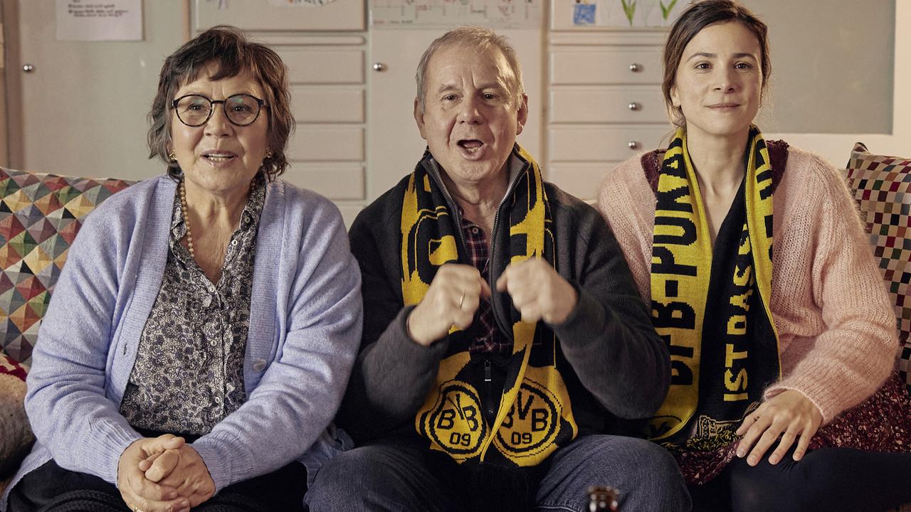 © ORF/Pro7/LEONINE Studios Zwei Frauen und ein Mann in ihrer Mitte sitzen auf einem Sofa und tragen schwarz-gelbe Borussia-Dortmund-Schals und schauen sich die TV-Übertragung eines Fußballspiels an