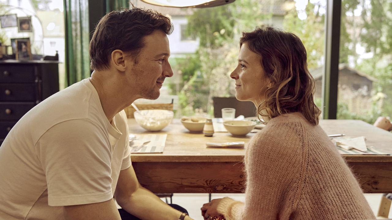 © ORF/Pro7/LEONINE Studios Ein Mann und eine Frau sitzen sich an einem gedeckten Holztisch gegenüber und schauen sich lächelnd und freundlich an