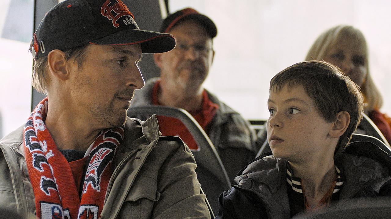 © ORF/Pro7/LEONINE Studios Mehrere Personen sitzen in einem Bus, ein Mann im Vordergrund trägt einen rot-weißen Fan-Schal und eine Kappe und schaut seinen neben ihm sitzenden Sohn an
