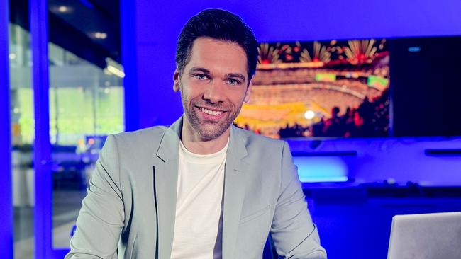 © ORF Philipp Maschl sitzt in einem modern beleuchteten Studio vor einem Laptop, während im Hintergrund auf einem Screen ein Stadion mit Feuerwerk zu sehen ist.