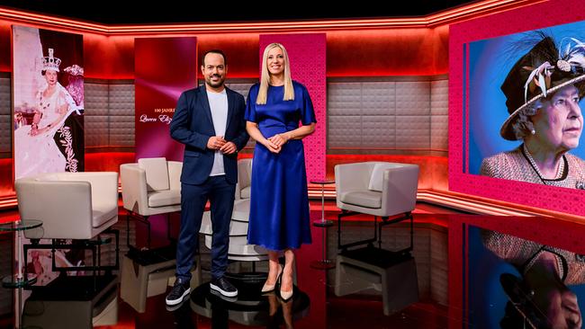 Patrick Budgen und Susanne Höggerl stehen in einem modern ausgeleuchteten TV‑Studio zwischen Sesseln und großen Porträtbildern von Queen Elizabeth II.