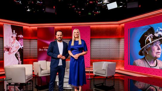 Patrick Budgen und Susanne Höggerl stehen in einem modern ausgeleuchteten TV‑Studio zwischen Sesseln und großen Porträtbildern von Queen Elizabeth II.