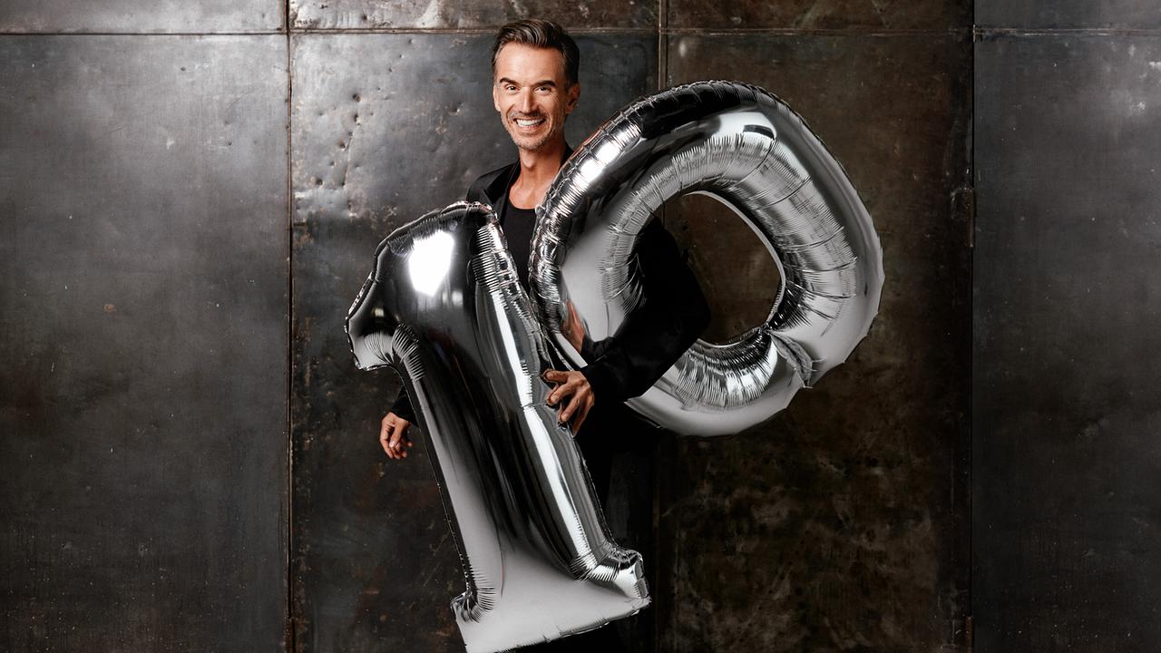 © ORF/ARD/Frank Zauritz Florian Silbereisen präsentiert "10 Jahre Schlagerbooom" und hält vor einer dunklen Wand große silberne Folienballons in Form der Zahl 10.
