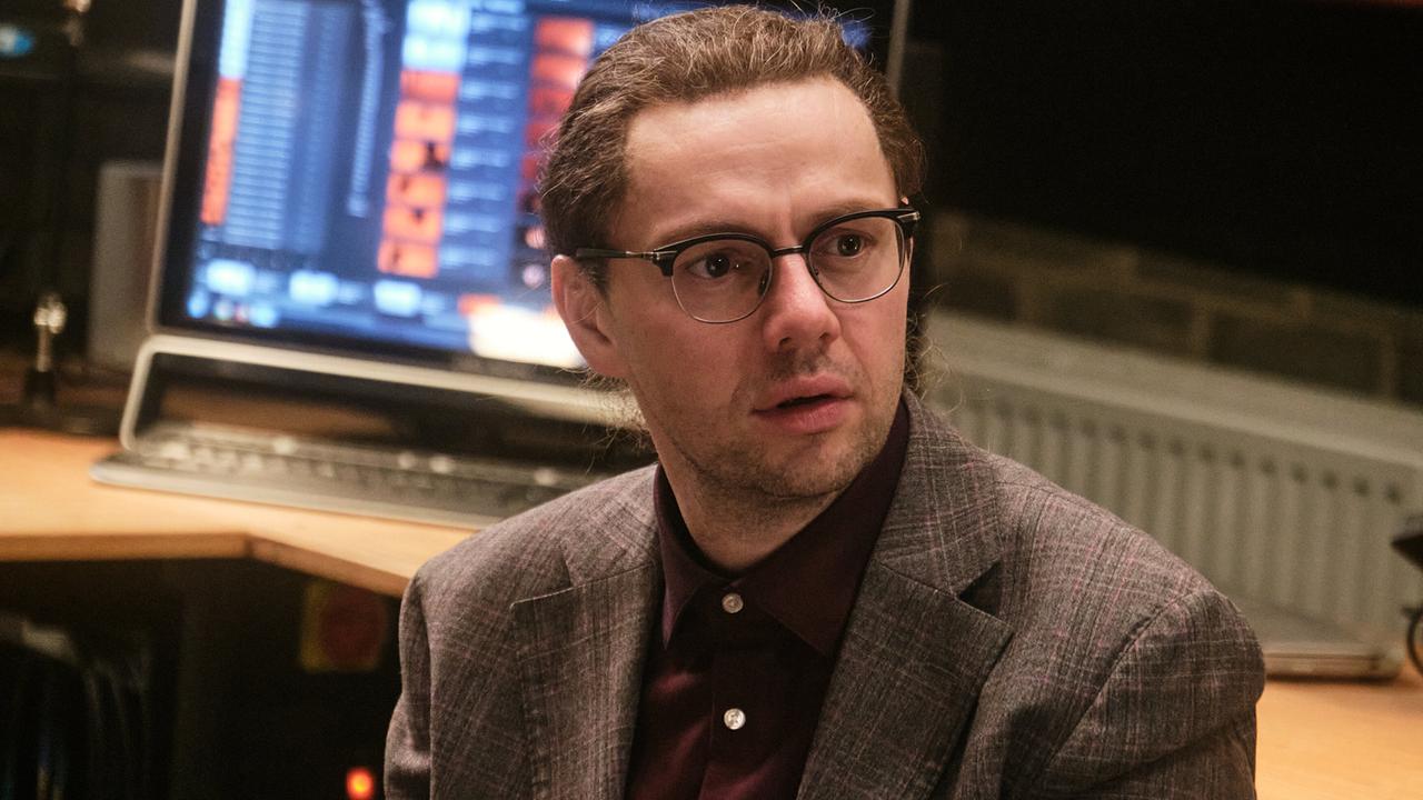 Harry Mangold sitzt verkehrt an einem Schreibtisch mit einem Bildschirm im Hintergrund und blickt nach vorne.