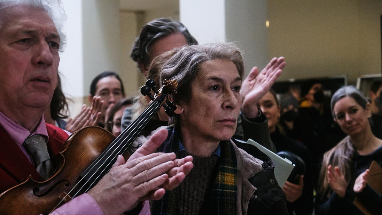 Frau Markowski steht ernst inmitten applaudierender Menschen, im Vordergrund hält ein Mann eine Geige.