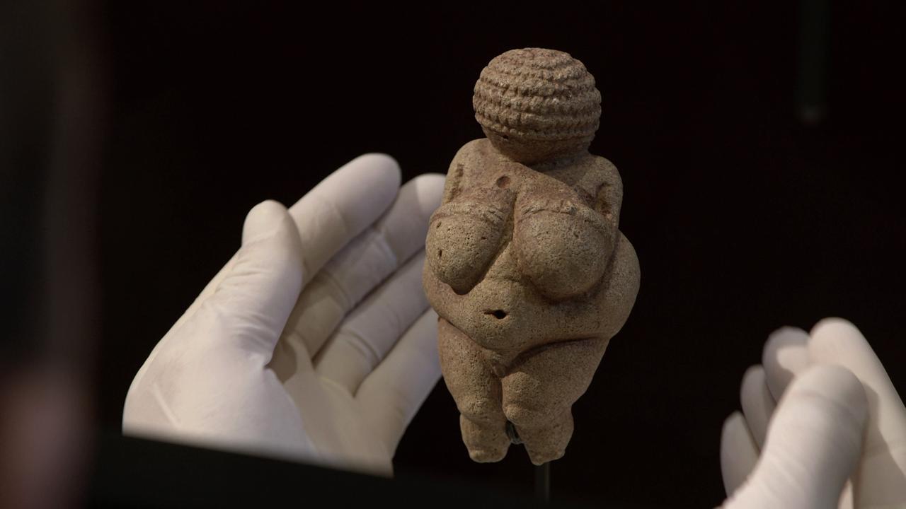 Venus von Willendorf vor schwarzem Hintergrund zwei in Plastikhandschuhen gehüllte Hände um sie