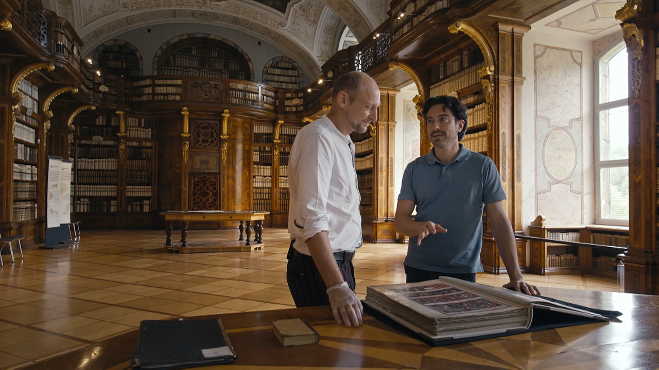 Schauspieler Stefano Bernardin und Kunsthistoriker Andreas Gamerith stehen mit einem großen, alten Buch in der Stiftsbibliothek Zwettl