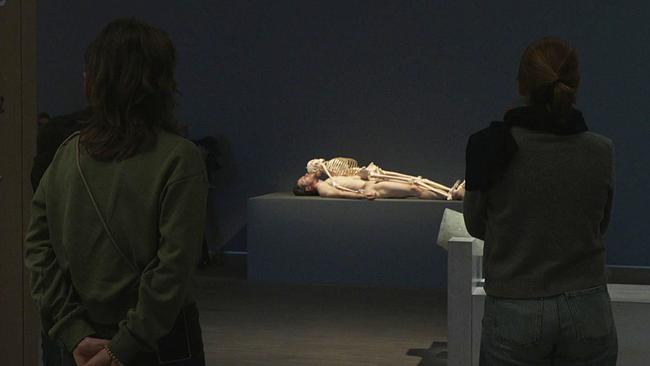 Ausstellungsansicht "Marina Abramović" - Besucher*innen vor liegendem nacktem Mann auf dem ein Skelett liegt