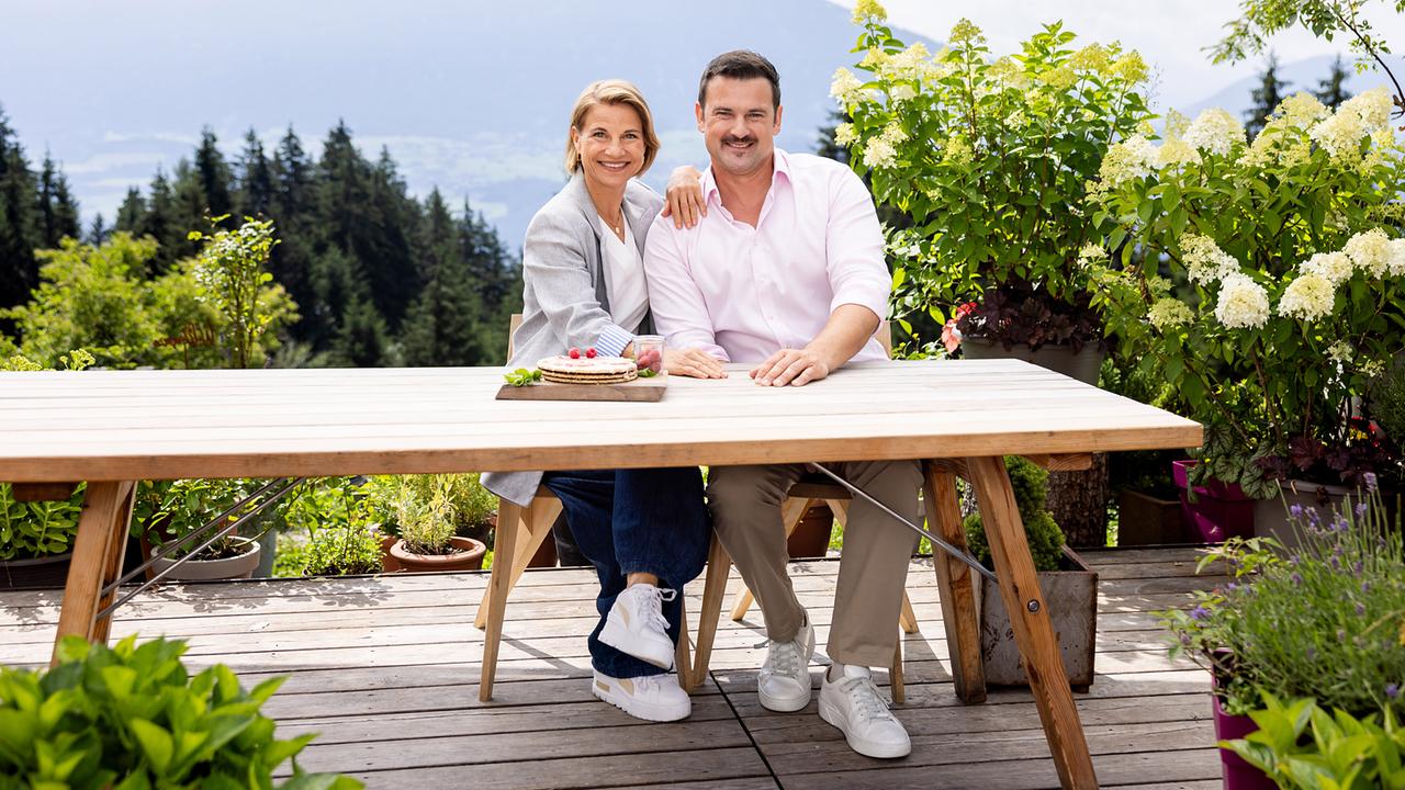 Das Moderations-Duo von "2 in Tirol" Kristina Sprenger und Sigi Fink sitzt an einem Holztisch auf einer Terrasse auf der Umbrüggler Alm.