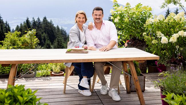 Das Moderations-Duo von "2 in Tirol" Kristina Sprenger und Sigi Fink sitzt an einem Holztisch auf einer Terrasse auf der Umbrüggler Alm.