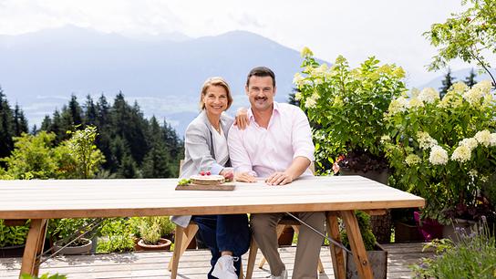 Das Moderations-Duo von "2 in Tirol" Kristina Sprenger und Sigi Fink sitzt an einem Holztisch auf einer Terrasse auf der Umbrüggler Alm.
