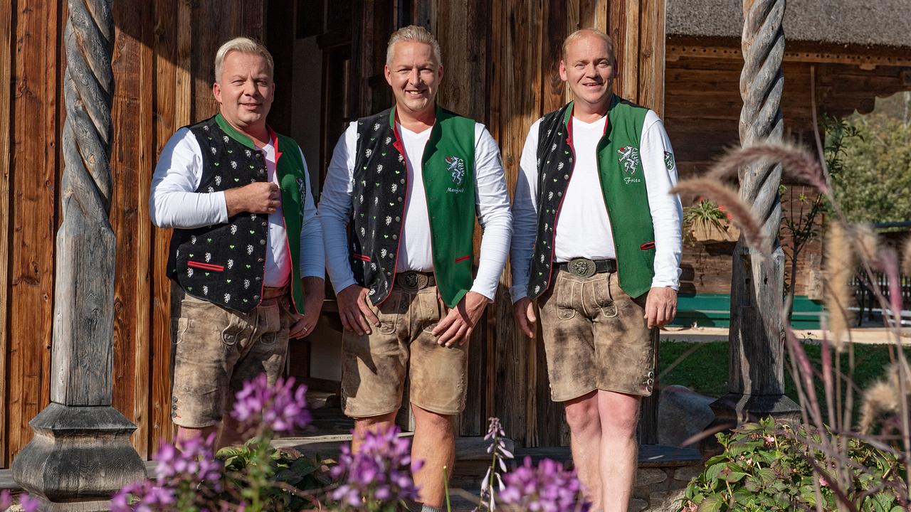Die Edlseer Andreas Doppelhofer, Manfred Maier und Fritz Kristoferitsch  in traditioneller Tracht stehen vor einem Holzhaus und posieren für ein Foto.