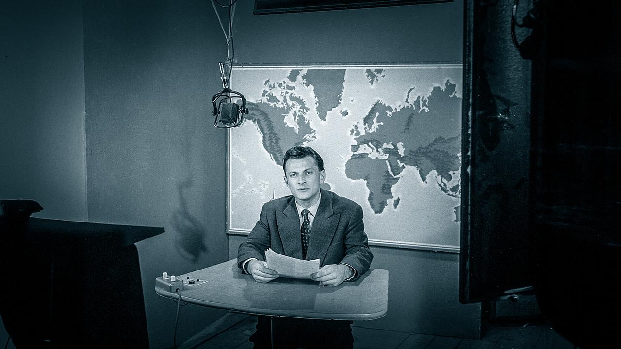 Herbert Kragora, der erste Sprecher der "Zeit im Bild" 1957, sitzt im "ZIB"-Studio am Nachrichtentisch vor einer Weltkarte mit einem Skript in der Hand.