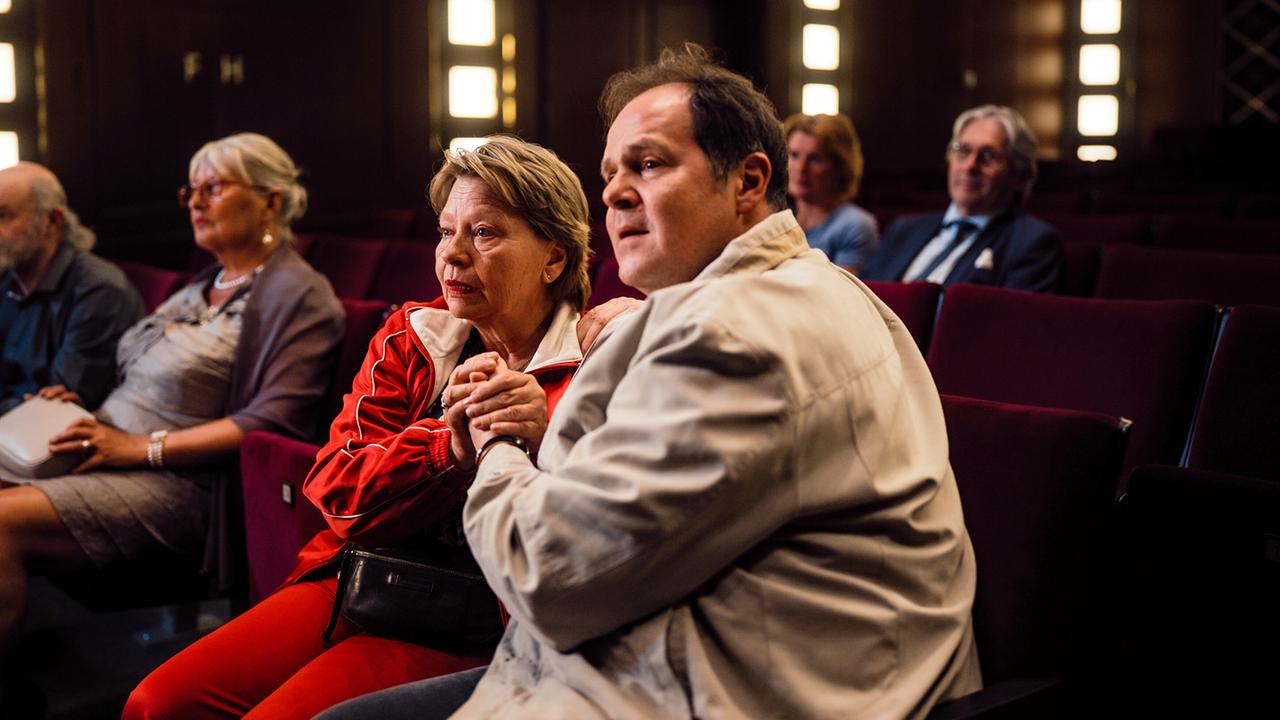 Toni und Ferdinand sitzen in einem Kinosaal und halten sich gespannt die Hände, im Hintergrund weitere Kinobesucher.