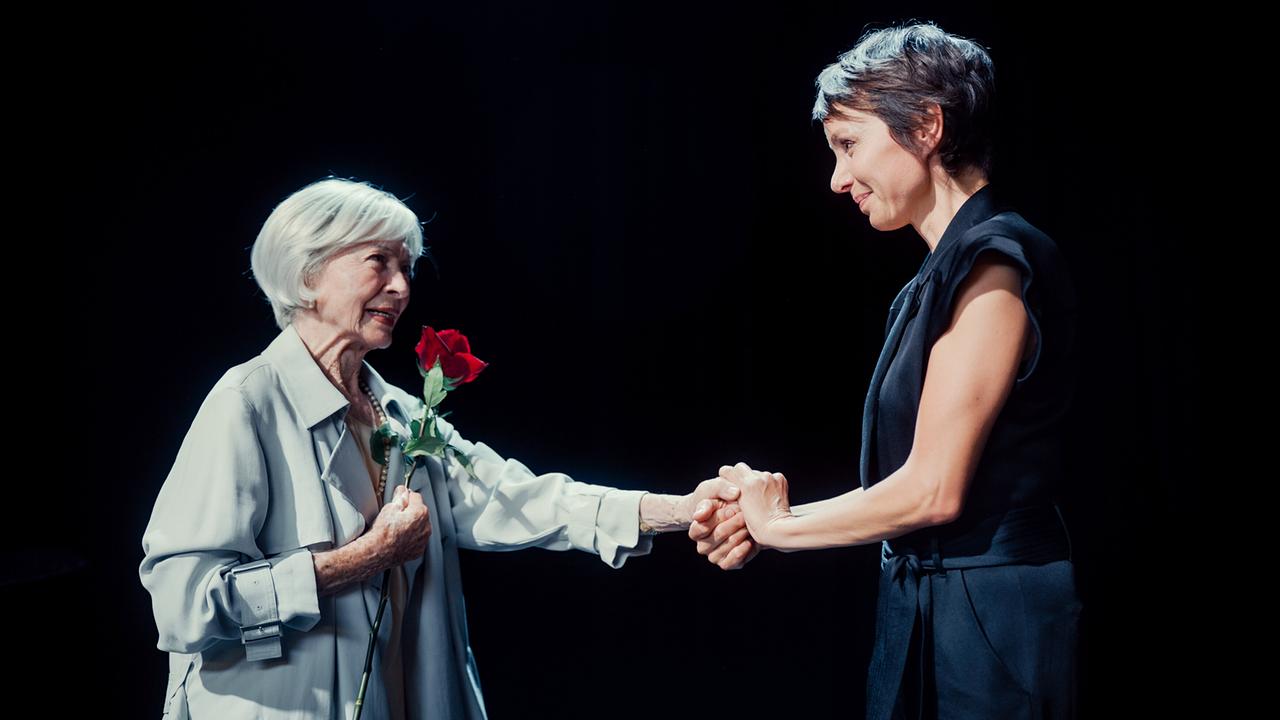 Helene mit einer Rose in der einen Hand drückt mit der anderen Hand die Hände von Thea, die ihr auf einer dunklen Bühne gegenüber steht.