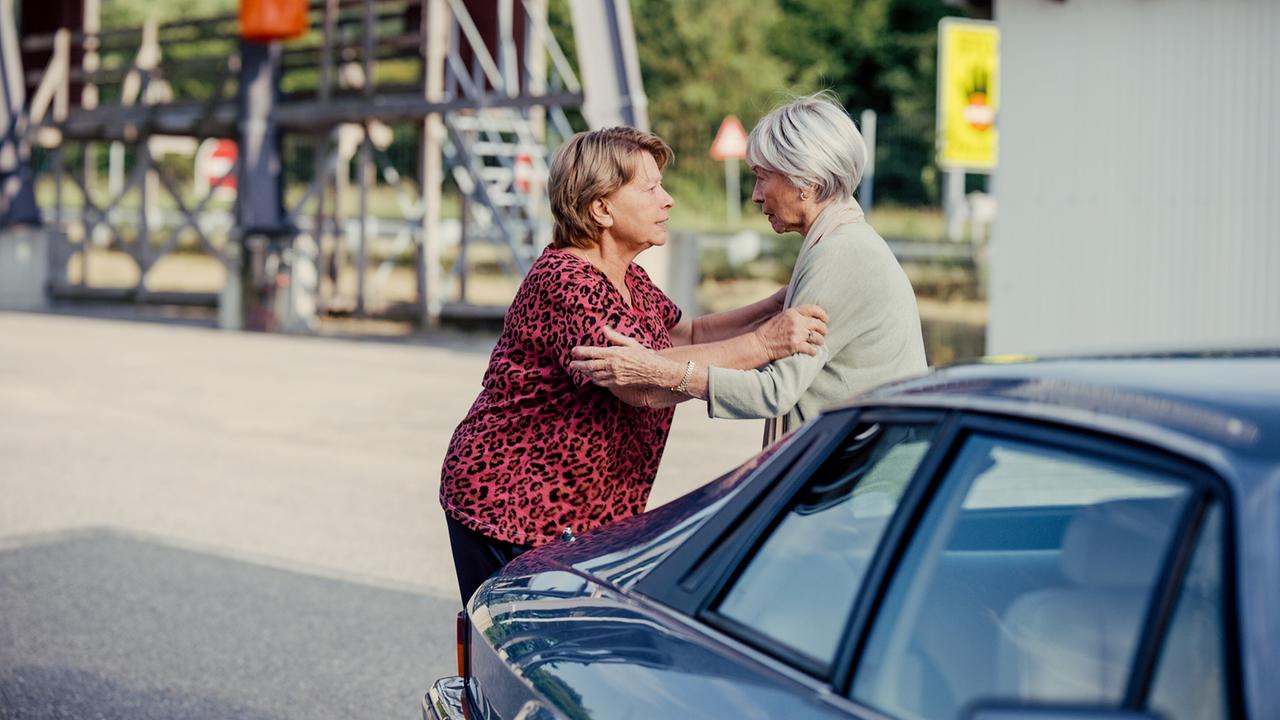 Toni und Helene umarmen sich neben einem Auto auf einem offenen Platz an der Grenze.
