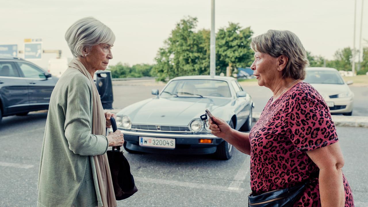 Helene und Toni, mit einem Autoschlüssel in der Hand, stehen vor Autos auf einem Parkplatz.