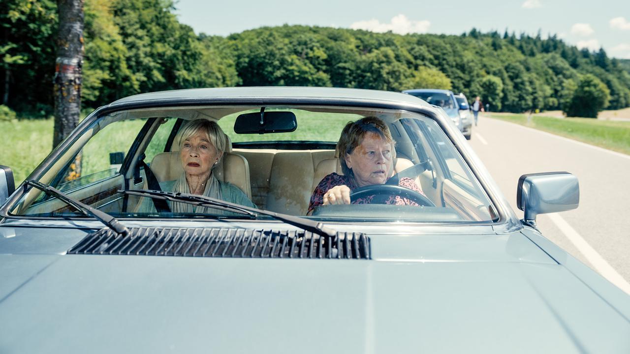 Helene und Toni am Steuer sitzen in einem Auto und fahren auf einer Landstraße durch eine grüne Landschaft, wobei Toni besorgt in den Rückspiegel blickt.