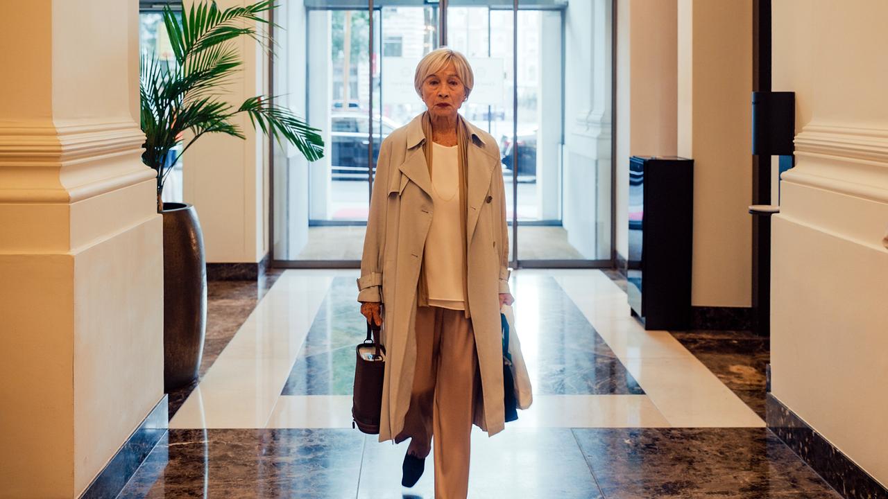 Helene geht mit Tasche und Mantel durch eine helle Lobby.