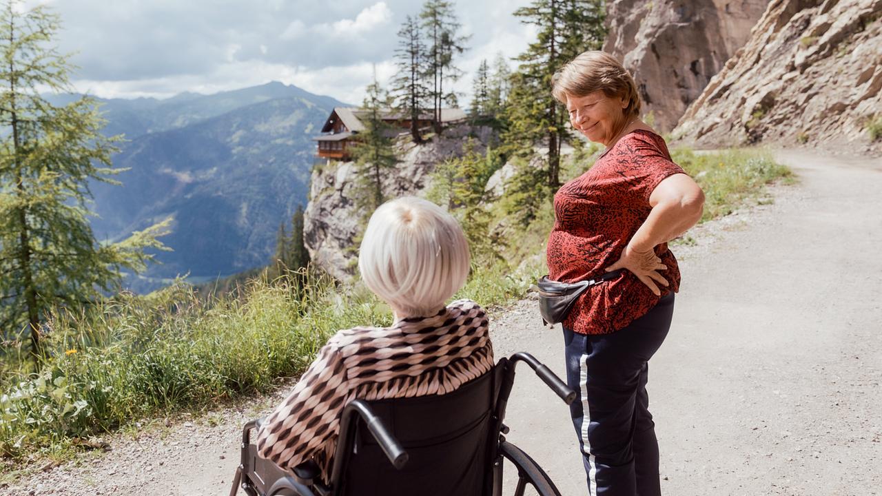 Helene im Rollstuhl und Toni daneben halten auf einem Bergweg mit Blick auf eine Berglandschaft.