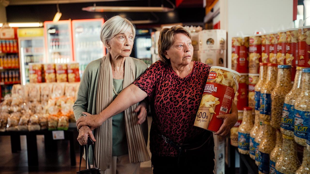 Helene und Toni gehen Arm in Arm durch einen Supermarkt, eine trägt eine 400-Gramm-Snackdose.