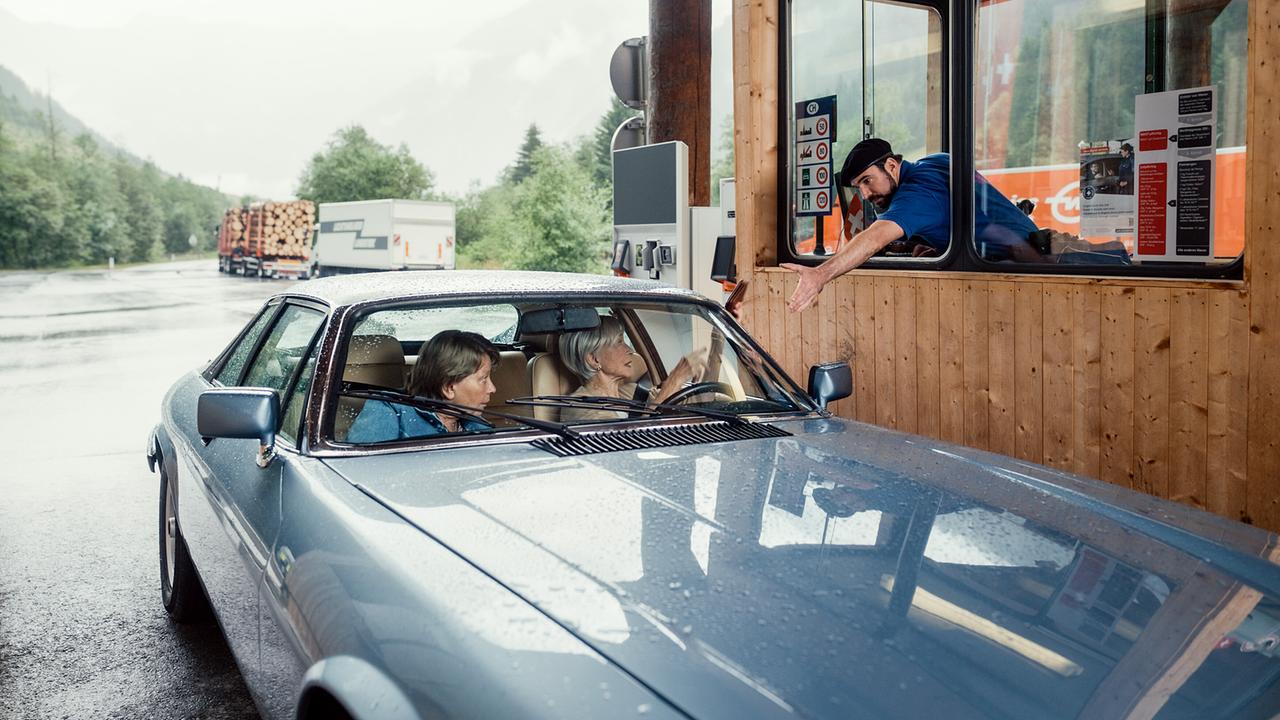 Toni und Helene im Auto an einer Grenzstation reichen aus dem geöffneten Fenster etwas dem Grenzbeamten, der in der Station sitzt.