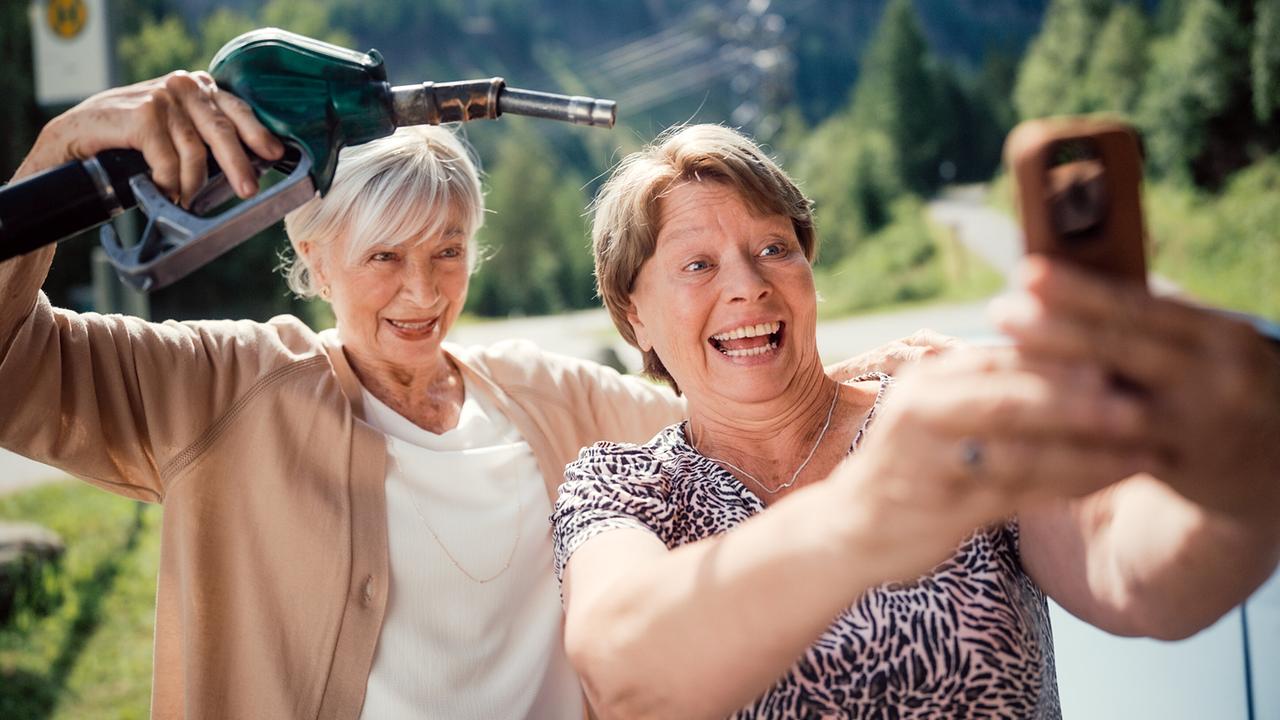 Helene und Toni machen in einer grünen Umgebung ein Selfie, während Helene scherzhaft eine Zapfpistole hochhält.