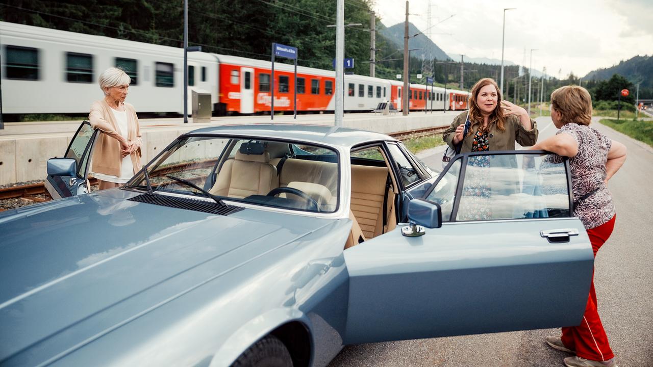 Helene und Toni stehen vor einer Autostopperin neben einem Auto an einem Bahnhof, im Hintergrund fährt ein Zug vorbei.