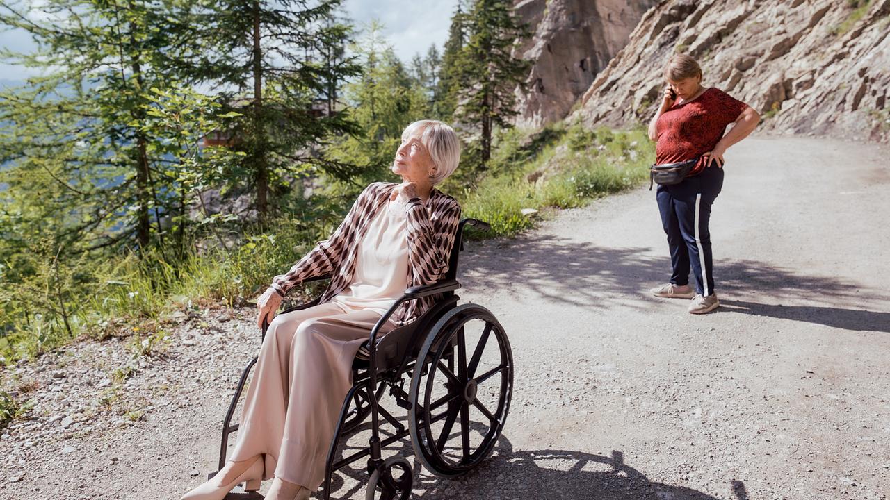 Helene sitzt im Rollstuhl auf einem Bergweg, während Toni dahinter steht und am Smartphone telefoniert.