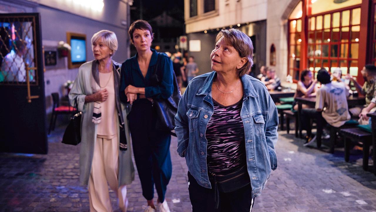Thea hält die Hand von Helene, während die beiden mit Toni nachts durch eine belebte Straße an Gastgärten vorbeigehen.