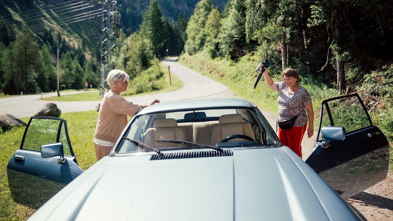 Helene und Toni stehen neben einem Auto an einer Bergstraße, eine hält eine lose Zapfpistole hoch.