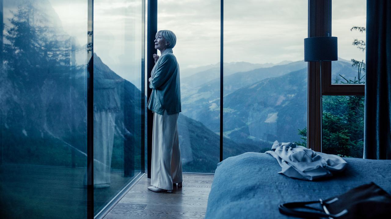 Helene blickt aus einem großen Fenster eines Hotelzimmers auf eine Berglandschaft.