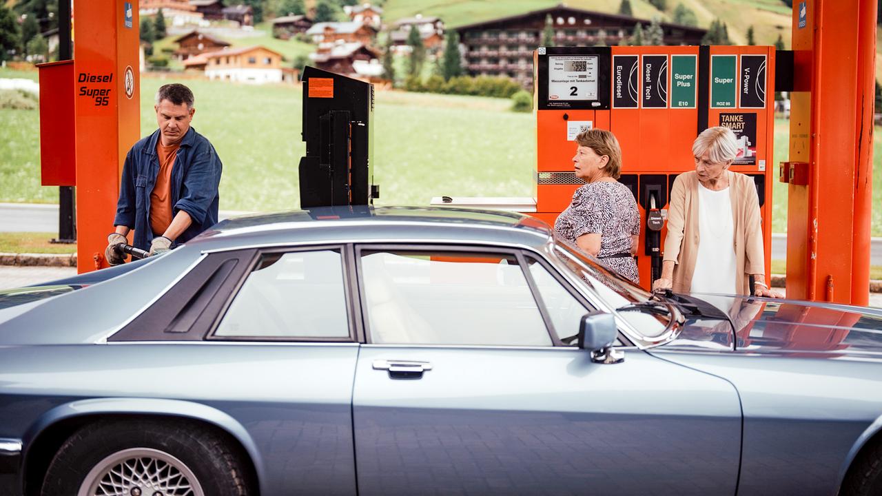 Ein Tankstellenbesitzer betankt ein Auto betankt vor alpiner Kulisse, Toni und Helene stehen daneben vor den Zapfsäulen.