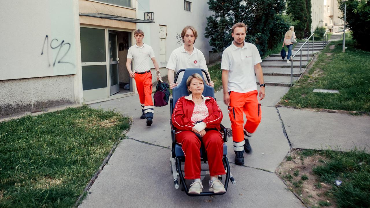 Sanitäter schieben Toni im Rollstuhl über einen Gehweg vor einem Wohnhaus.