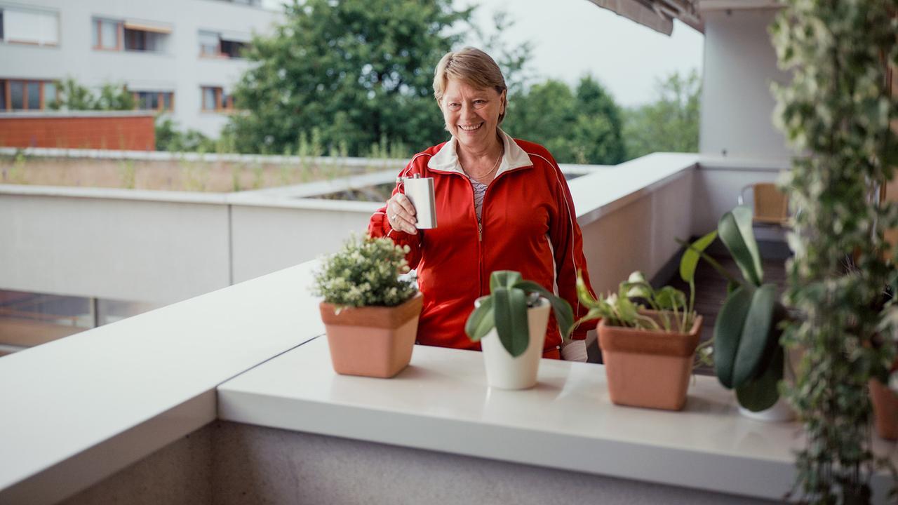 Toni steht auf einem Balkon zwischen Pflanzen und hält lächelnd einen Flachmann in der Hand.