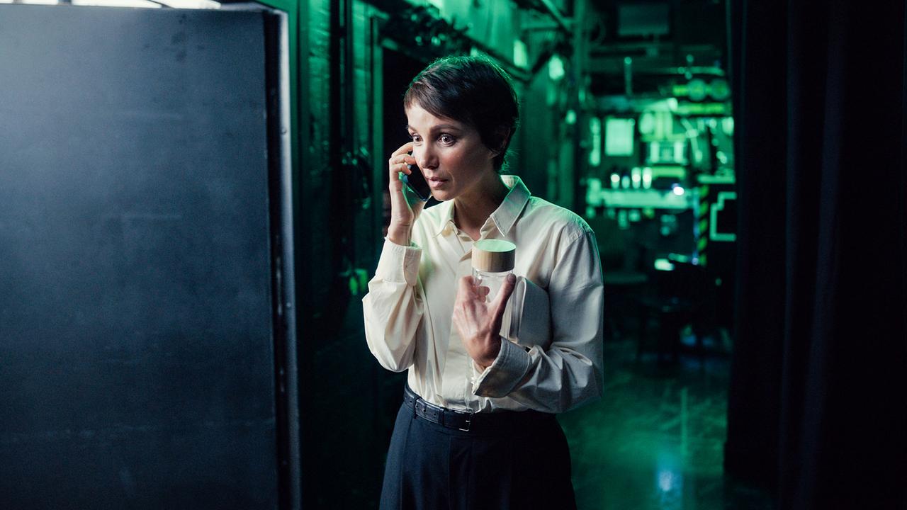 Thea telefoniert in einem dunklen Technikraum und hält dabei eine Glasflasche in der Hand.