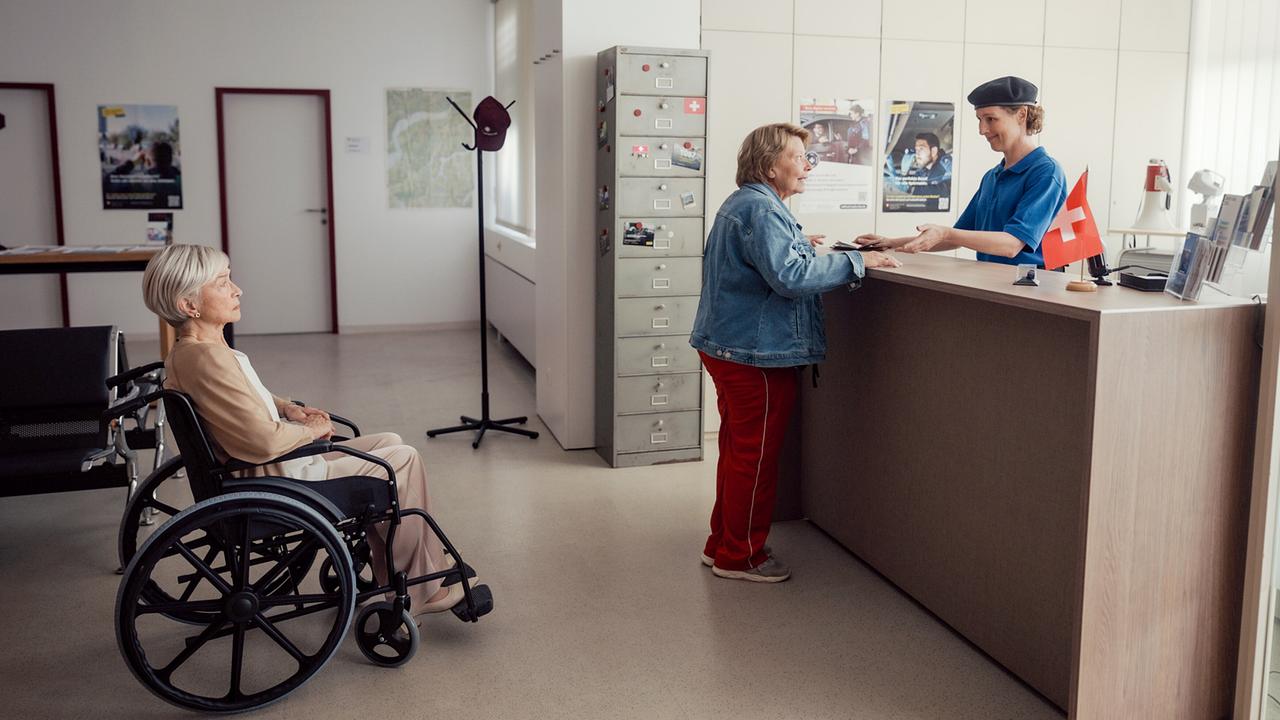 In einem hellen Amtsraum spricht Toni mit der Grenzbeamtin am Schalter, während Helene im Rollstuhl dahinter zusieht