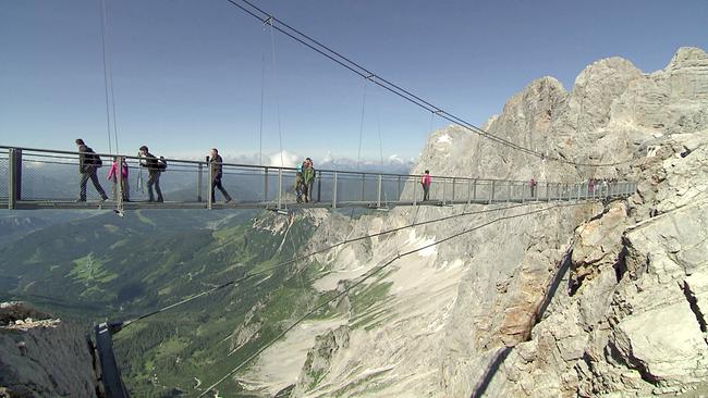 © ORF Wander überqueren die Dachstein Hängebrücke mit Blick auf das Tal und felsige Bergwände.