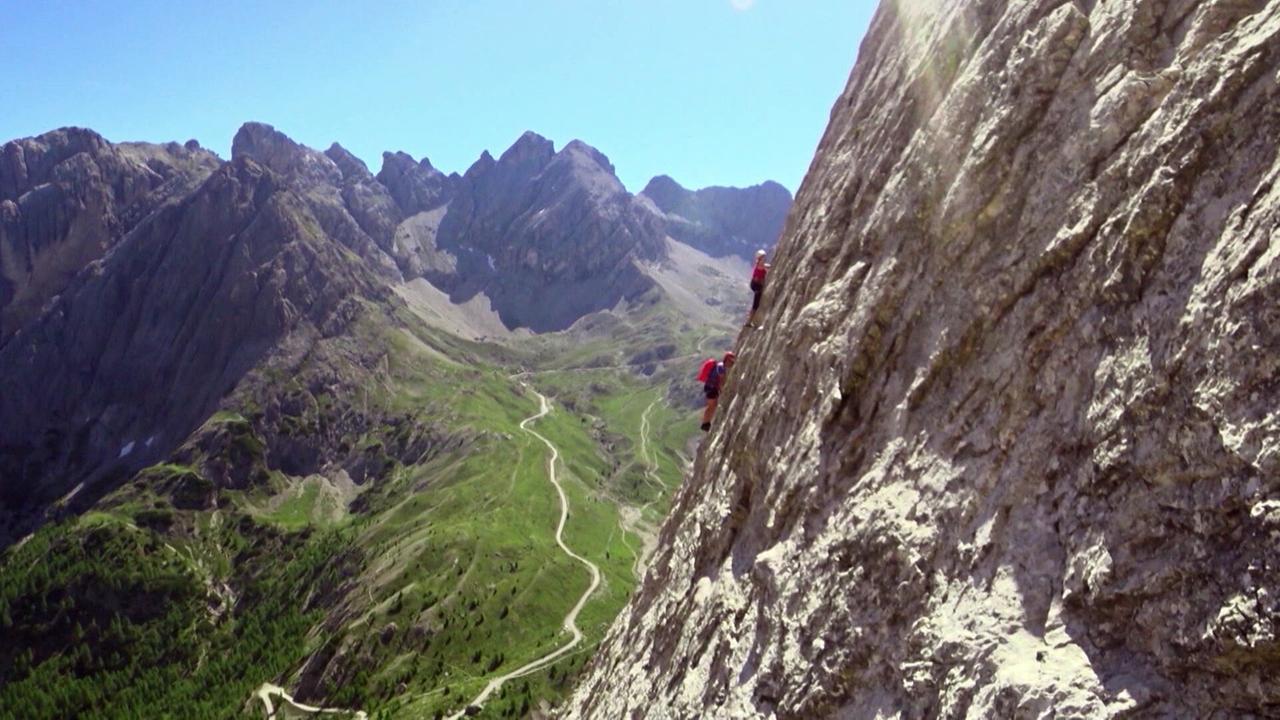 © ORF Bergsteiger klettern an steiler Felswand vor grünem Hochtal und Gipfeln der Lienzer Dolomiten.