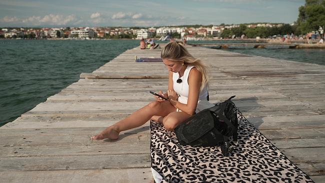 © ORF Julijana sitz auf einem Tuch auf einem Steg am Meer, sie schaut in ihr Handy, im Hintergrund das Meer und die Küste