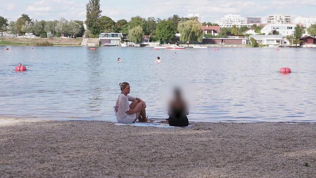 Strand eines Schwimmbades an der Donau  - am Strand beim Wasser sitzen Redakteurin Marianne Pušić und eine Frau, die man von hinten sieht - am anderren Ufer Boote, Bäume und Gebäude 