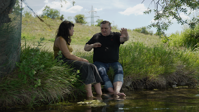 Redakteurin Tara Giahi sitzt mit Admir Hasanović an einem Flußuffer im Schatten eines Baumes, sie lassen die Füße ins Wasser baumeln