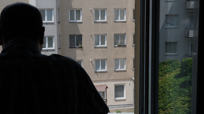 © ORF Ein Mann steht an einem Fenster im Schatten und sieht hinaus auf die Häuser auf der anderen Straßenseite