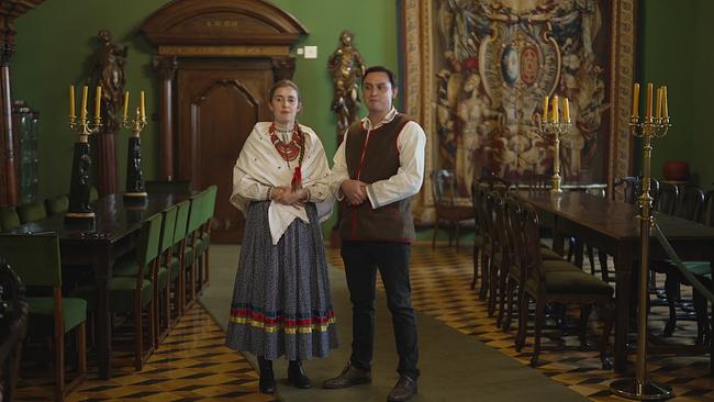 Junges Paar in Volkstracht in einem Raum mit 2 großen Tischen mit Kerzenleuchtern darauf - im Hintergrund eine alte Holztüre und ein Gobelin an der Wand