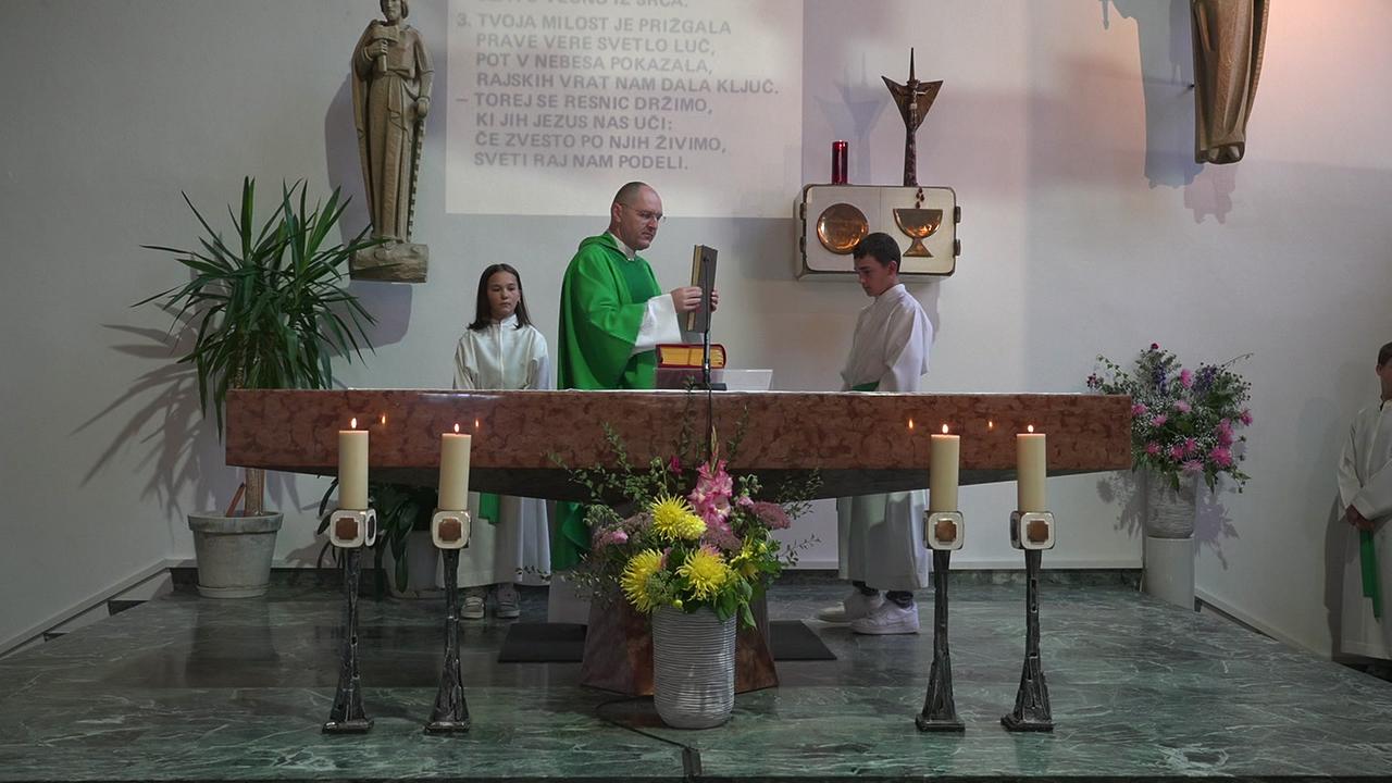 Altar in einer Kirche, dahinter steht ein Priester bei der Wandlung, flankiert von 2 Ministranten, eine slowenischsprachige Inschrift ist an der Wand zu sehen