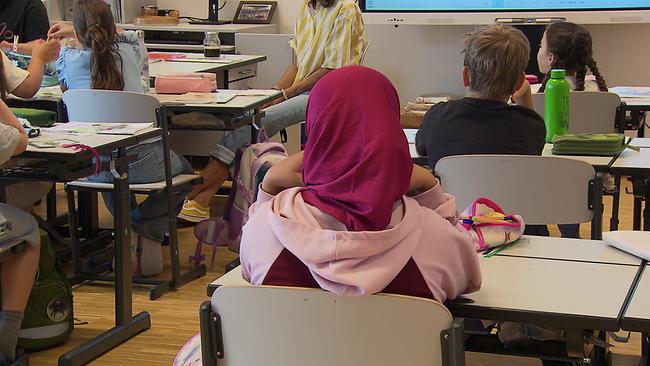 Mädchen mit Kopftuch und weitere Kinder sitzen in einer Schulklasse an ihren Schreibtischen - sie schauen zur Tafel, man sieht sie von hinten