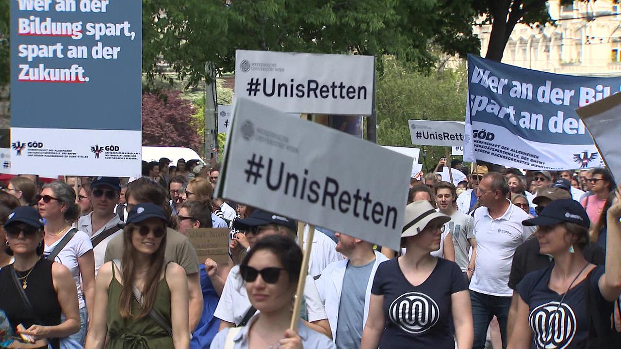 Gruppe protestierender Studenten - einige halten Schilder in die Höhe, auf denen folgendes steht "Wer an der Bildung spart, spart an der Zukunft" und "#Unis Retten"