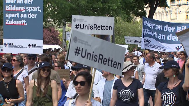 Gruppe protestierender Studenten - einige halten Schilder in die Höhe, auf denen folgendes steht "Wer an der Bildung spart, spart an der Zukunft" und "#Unis Retten"